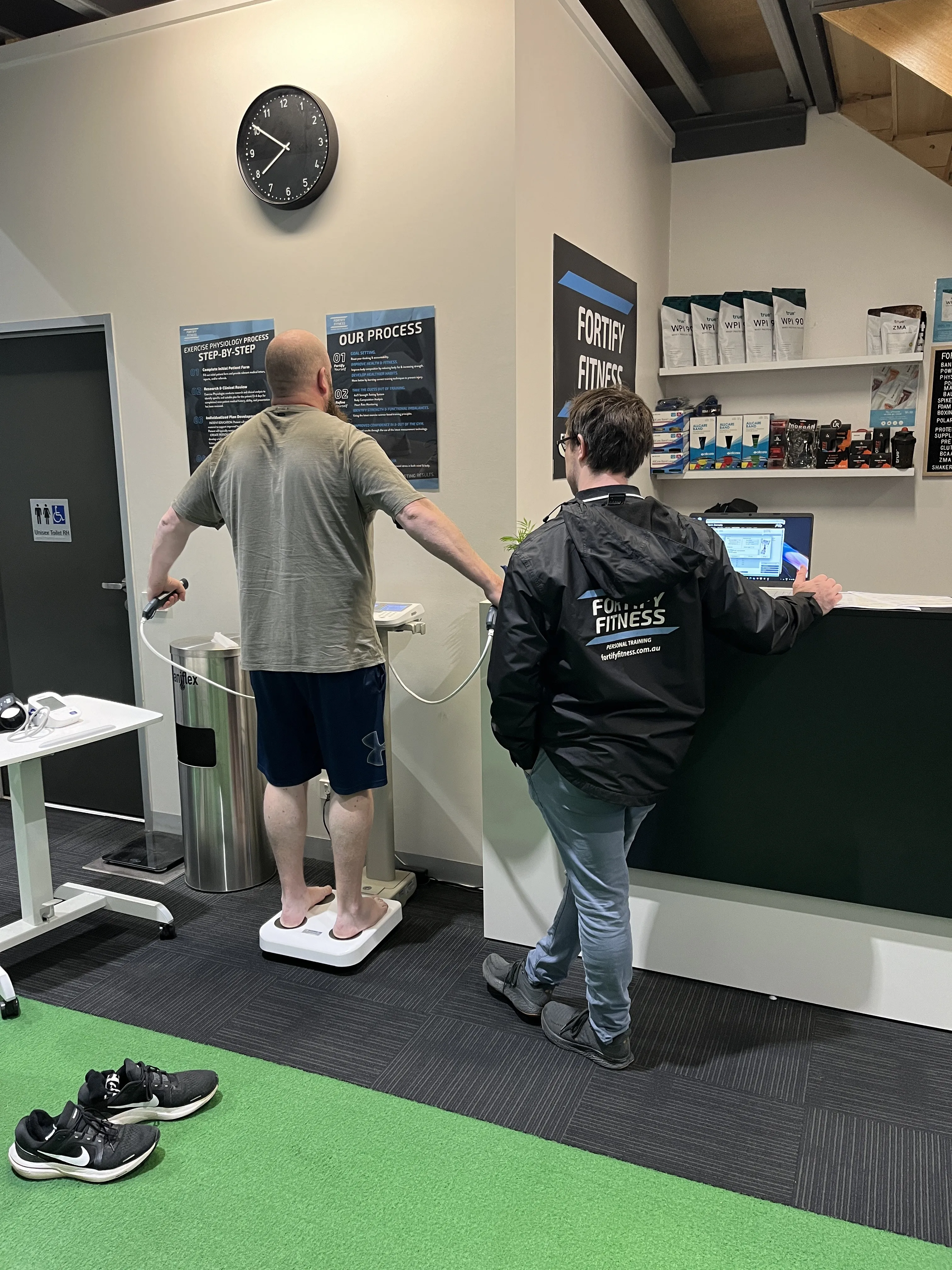 A man stands on a body composition scale while a staff member assists at a fitness center.